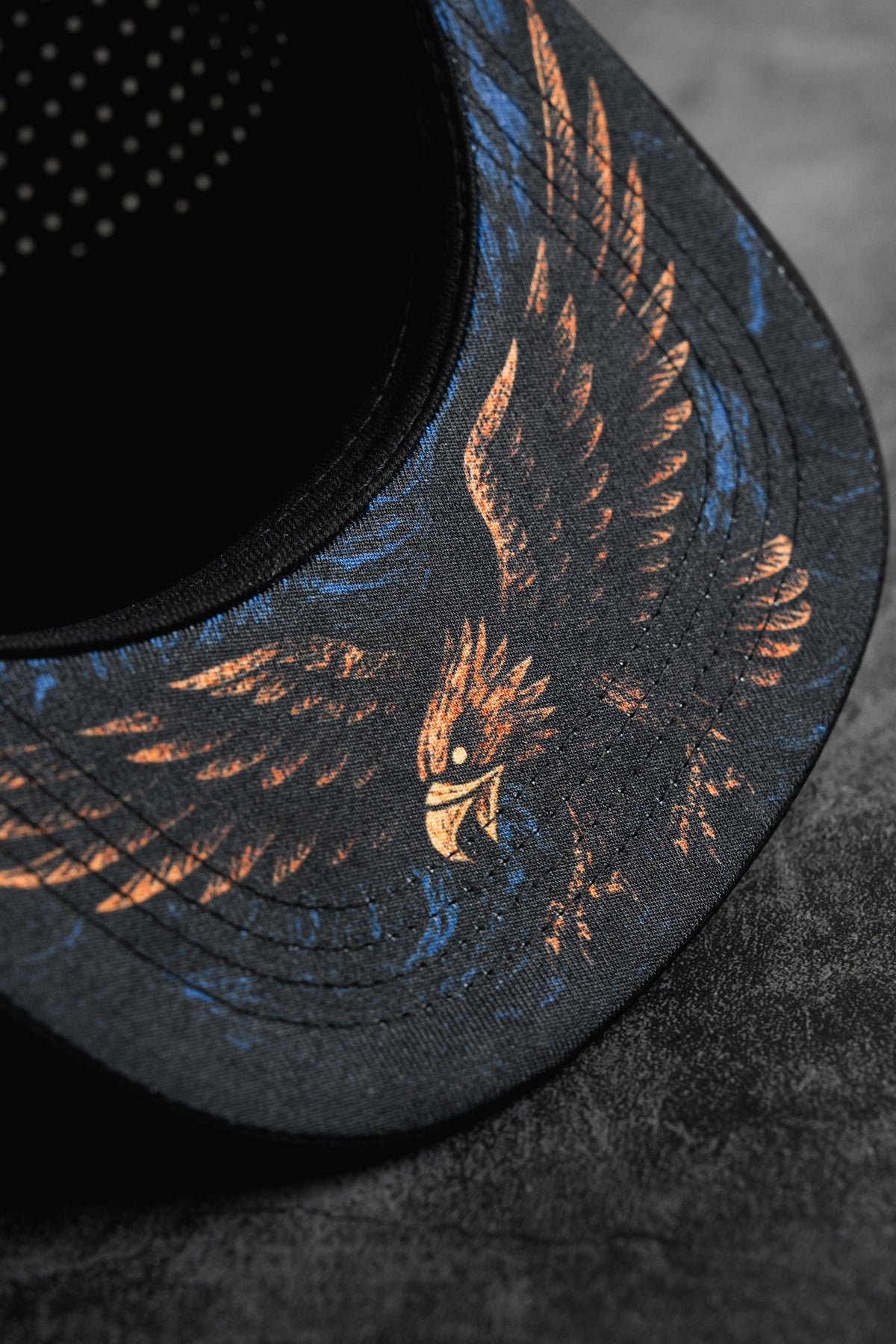 Close-up of the Relentless Betrayal Hold Steady Snapback, a premium black hat with an illustrated eagle in orange and blue on the brim, displayed on a textured dark gray surface.