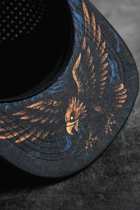 Close-up of the Relentless Betrayal Hold Steady Snapback, a premium black hat with an illustrated eagle in orange and blue on the brim, displayed on a textured dark gray surface.