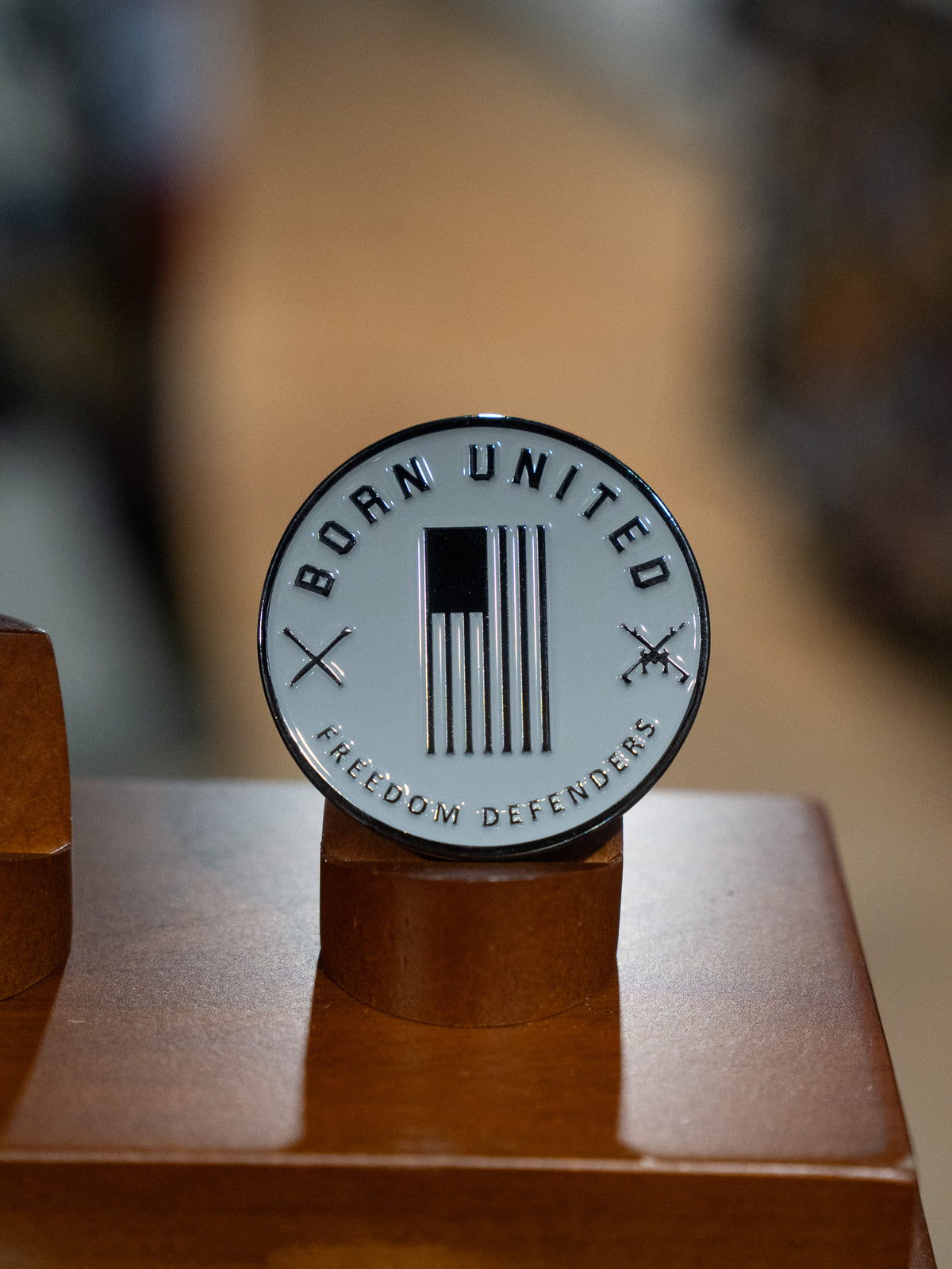 The Ain't Goofin' Around Challenge Coin by Born United Private Label features a round black and white U.S. flag with crossed rifles, "BORN UNITED" at the top, and "FREEDOM DEFENDERS" at the bottom, displayed on a wooden stand.