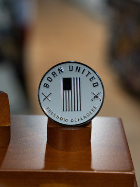 The Ain't Goofin' Around Challenge Coin by Born United Private Label features a round black and white U.S. flag with crossed rifles, "BORN UNITED" at the top, and "FREEDOM DEFENDERS" at the bottom, displayed on a wooden stand.