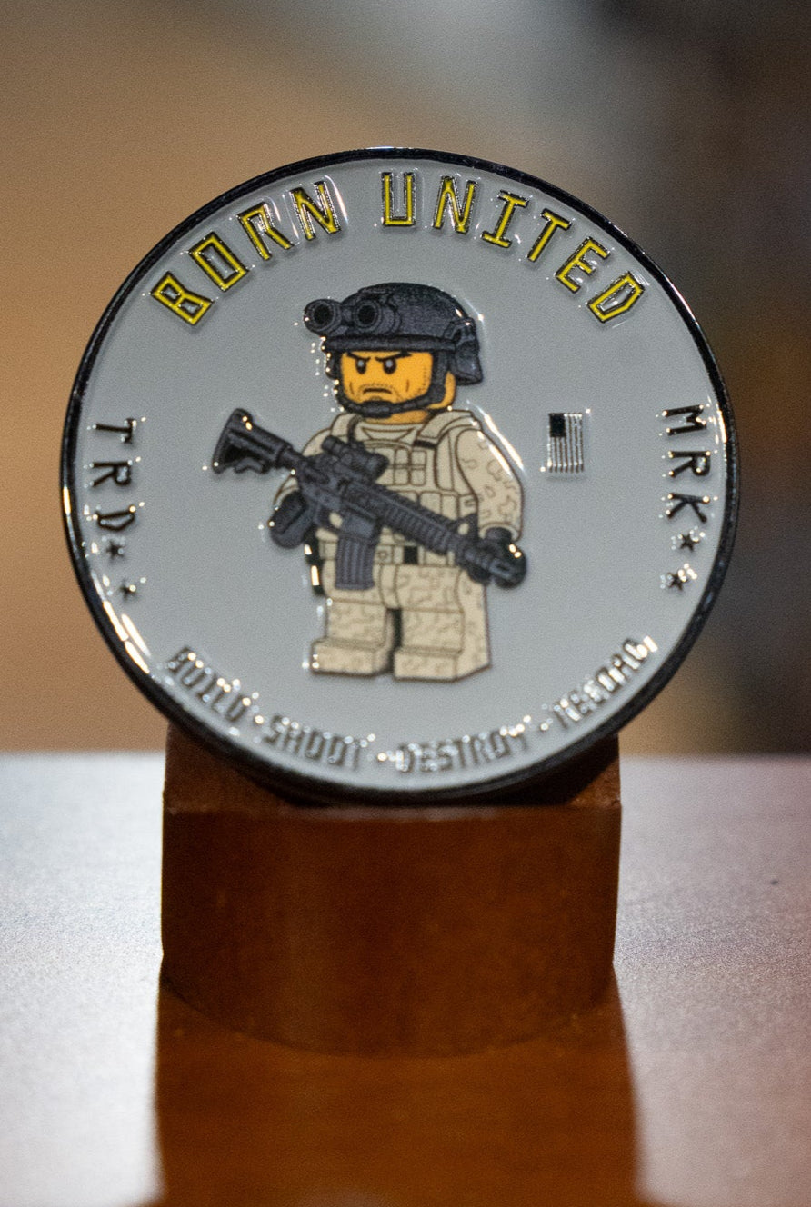 The Brick Operator Challenge Coin by Born United Private Label features a LEGO-style soldier with a rifle, helmet, and gear. "BORN UNITED" arcs above, "TRY" and "MRK" are at the sides, and "BUILD SHOOT DESTROY REBUILD" appears at the bottom.