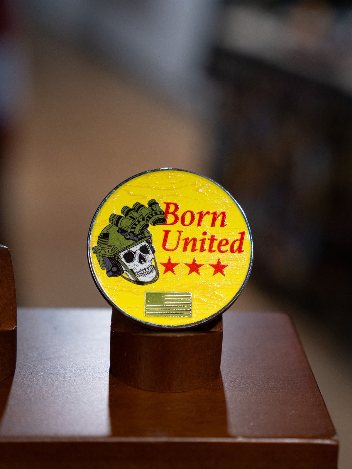 The Brass Pros Challenge Coin by Born United Private Label is a round yellow brass coin featuring a skull in a military helmet with night vision goggles, three red stars, and the words "Born United," displayed upright on a brown stand.