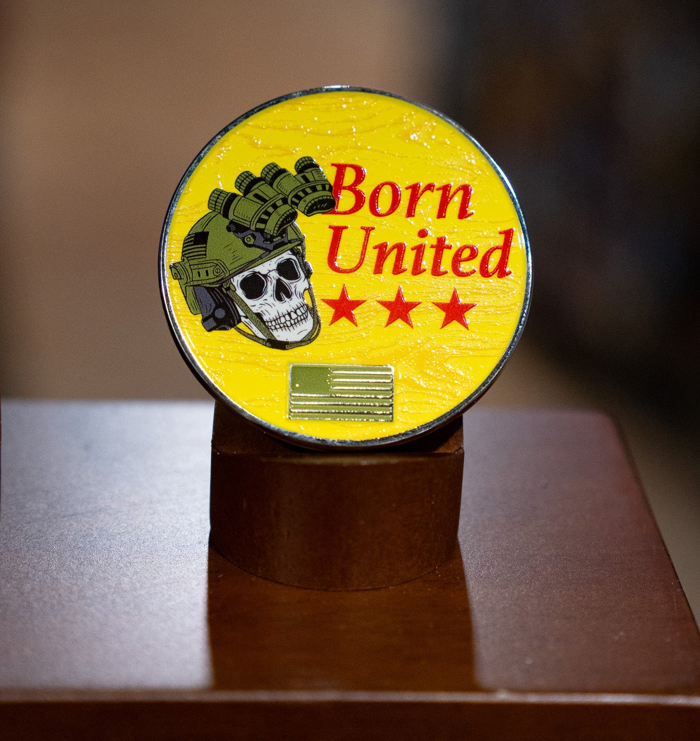 The Brass Pros Challenge Coin by Born United Private Label is a round yellow brass coin featuring a skull in a military helmet with night vision goggles, three red stars, and the words "Born United," displayed upright on a brown stand.