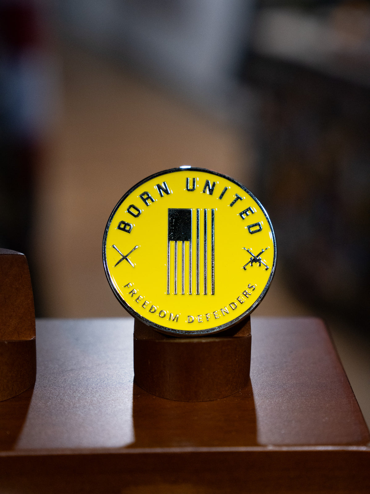 The Brass Pros Challenge Coin by Born United Private Label features a round yellow brass badge with a black vertical American flag, crossed rifles, and the words "BORN UNITED" and "FREEDOM DEFENDERS," displayed on a brown stand.