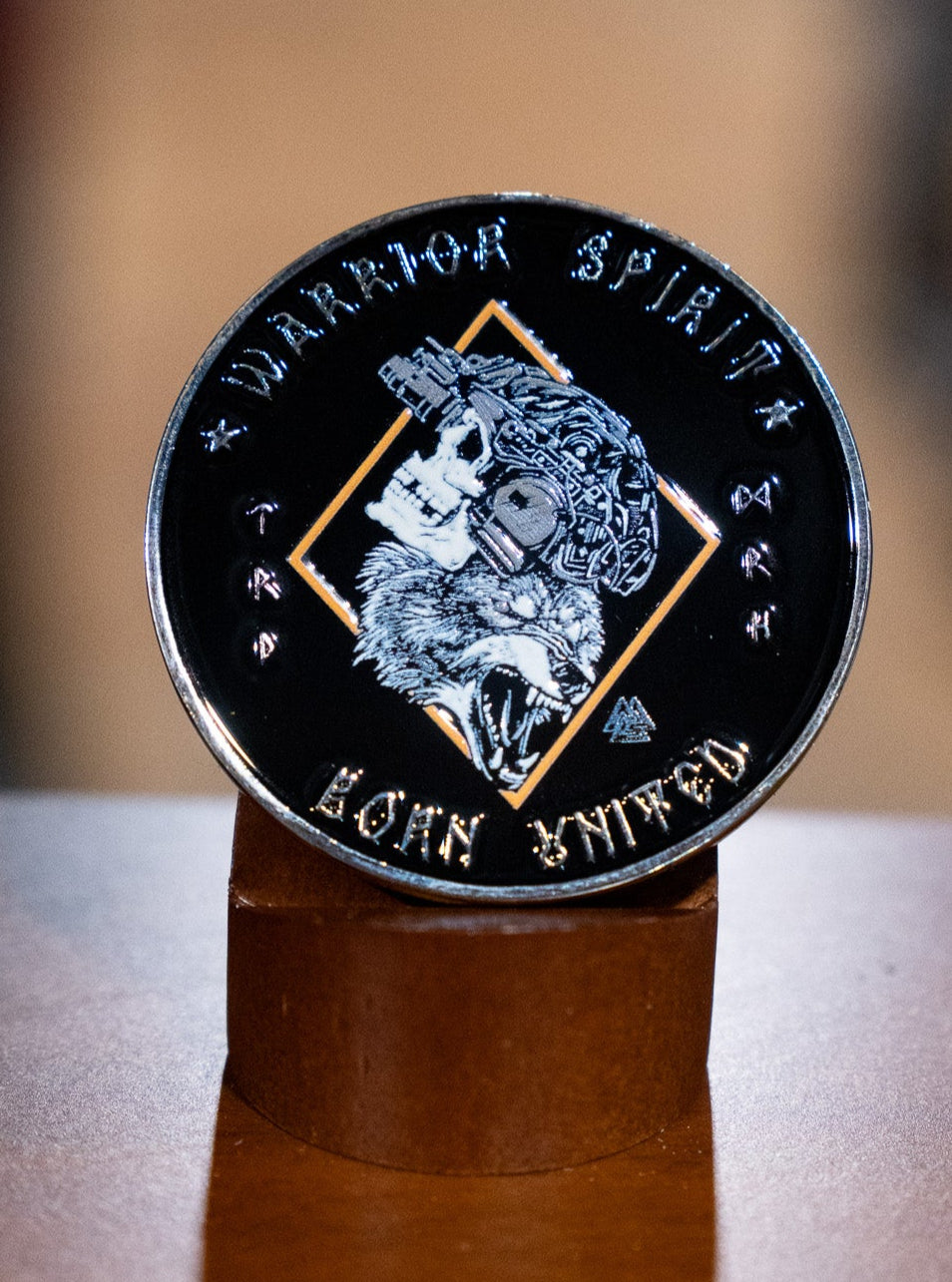 The Warrior Spirit Challenge Coin by Born United Private Label is a round black coin with a wolf’s head in a tactical helmet, "Warrior Spirit" inscribed at the top, and "BORN UNITED" at the bottom, displayed upright on a wooden stand.