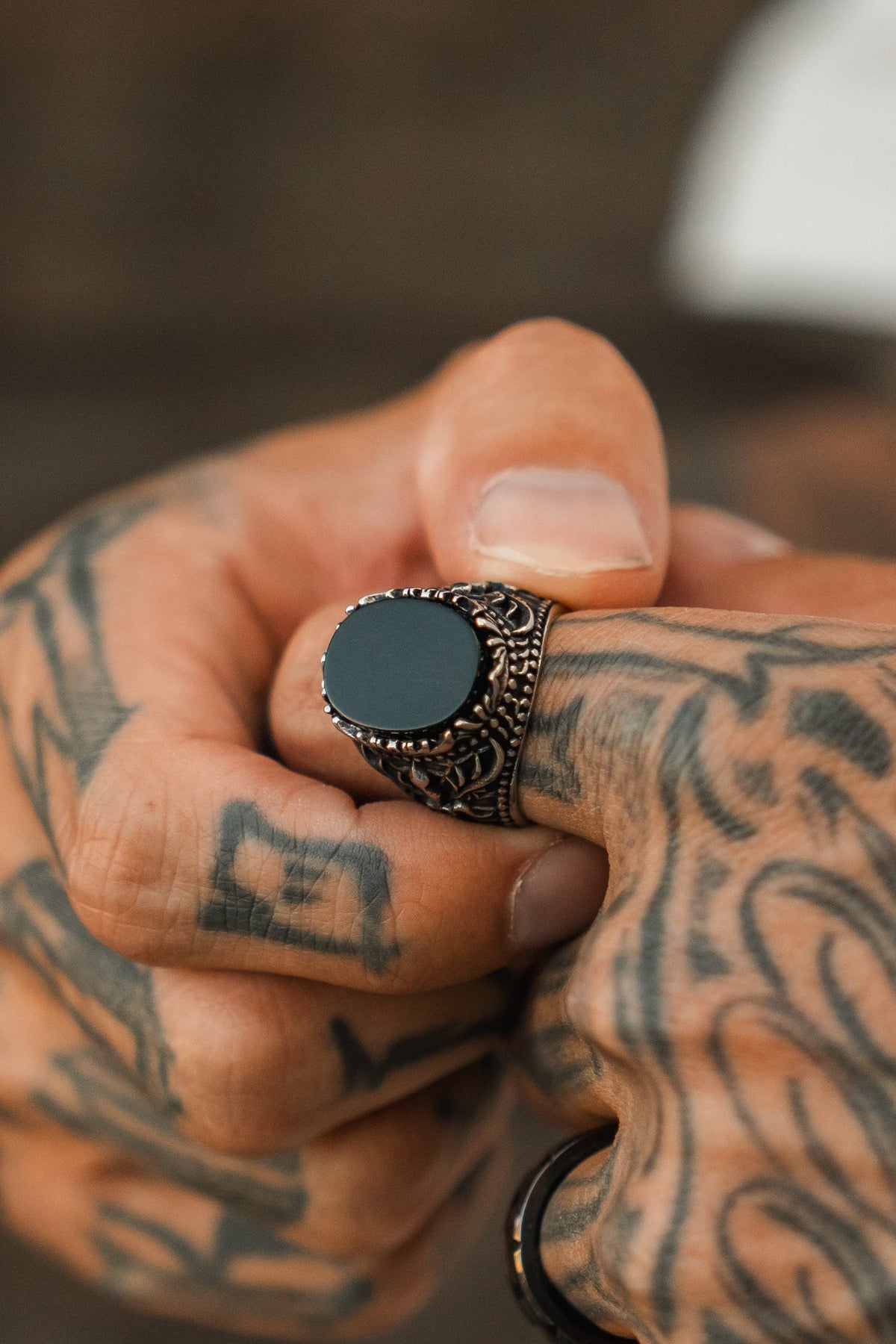 A tattooed hand wears The Dark Current Ring by Relentless Betrayal—a handcrafted men’s ring with a striking black stone and finish. The detailed black ink tattoos enhance the bold look, set against a softly blurred background.