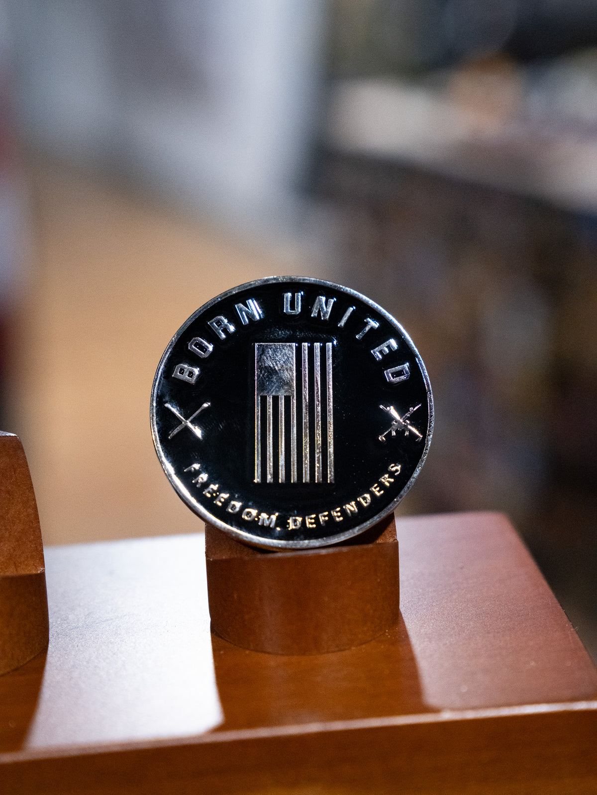 The Warrior Spirit Challenge Coin by Born United features a stylized American flag, “BORN UNITED” above, “FREEDOM DEFENDERS” below, and crossed rifles—symbolizing true warrior spirit—displayed on a wooden stand.