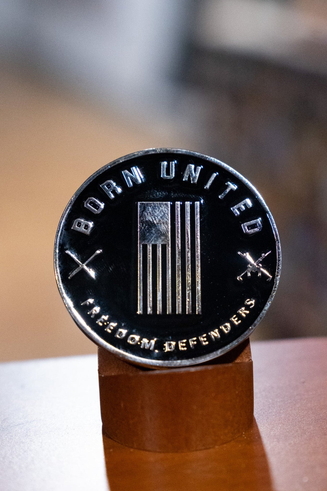 The Warrior Spirit Challenge Coin by Born United features a stylized American flag, “BORN UNITED” above, “FREEDOM DEFENDERS” below, and crossed rifles—symbolizing true warrior spirit—displayed on a wooden stand.