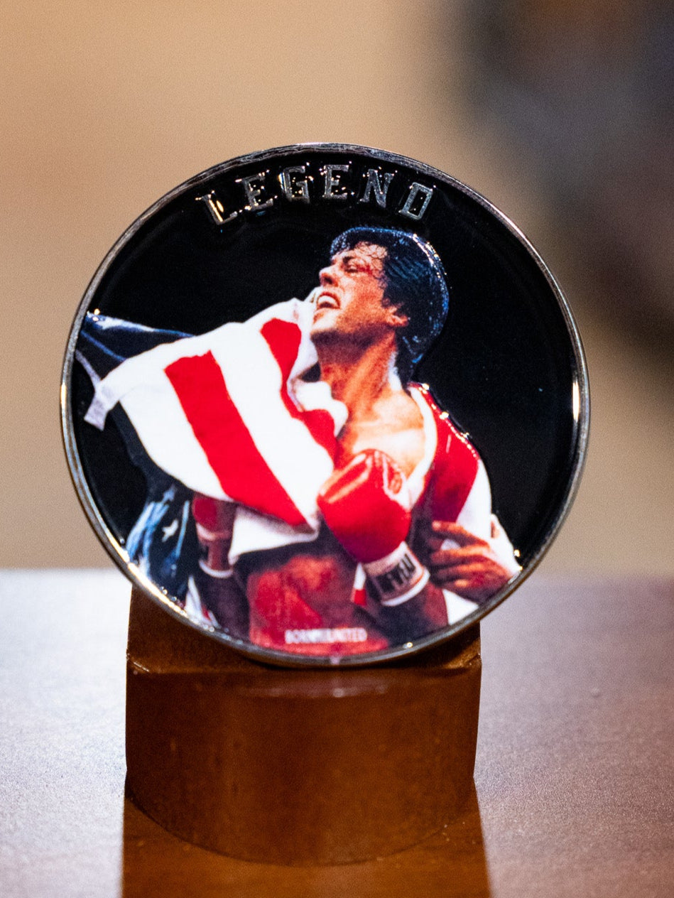 The Legend - Balboa Challenge Coin by Born United Private Label features the iconic boxer in red gloves holding an American flag, "LEGEND" above him, standing triumphantly on a brown stand.