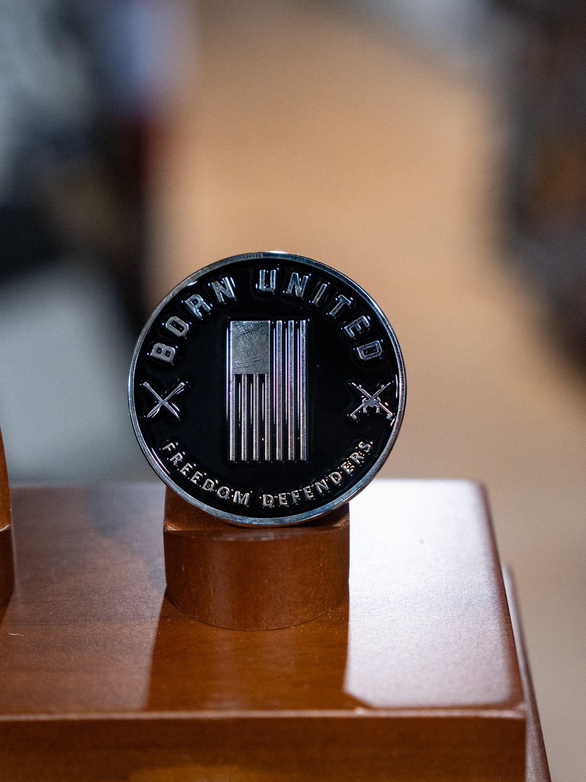 The Waking Lions Challenge Coin by Born United Private Label features an American flag, crossed rifles, and the words BORN UNITED and FREEDOM DEFENDER—a striking black collectible displayed on wood.