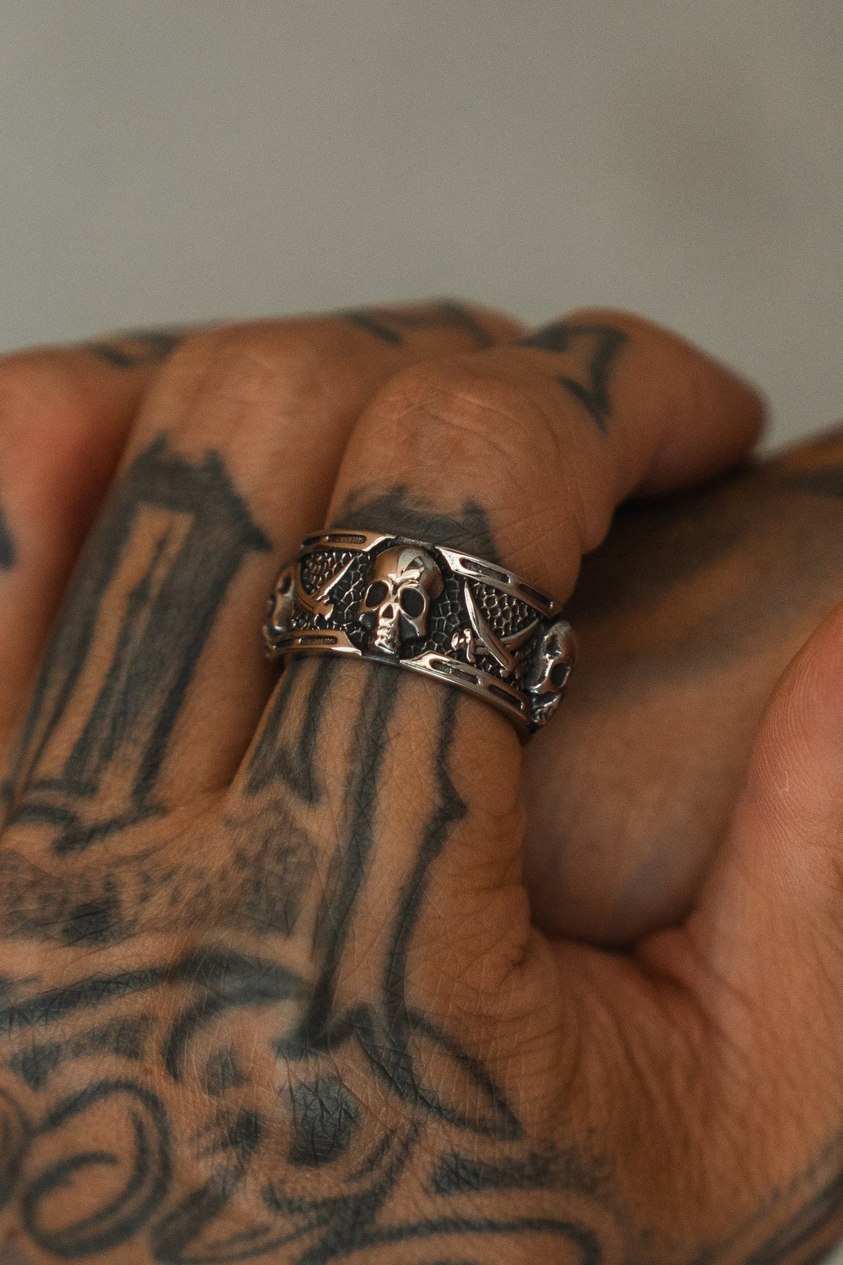 A close-up of a tattooed hand wearing the Relentless Betrayal Piracy Ring, featuring bold skull and wing details. The blurred background highlights the intricate tattoos and the standout ring design.