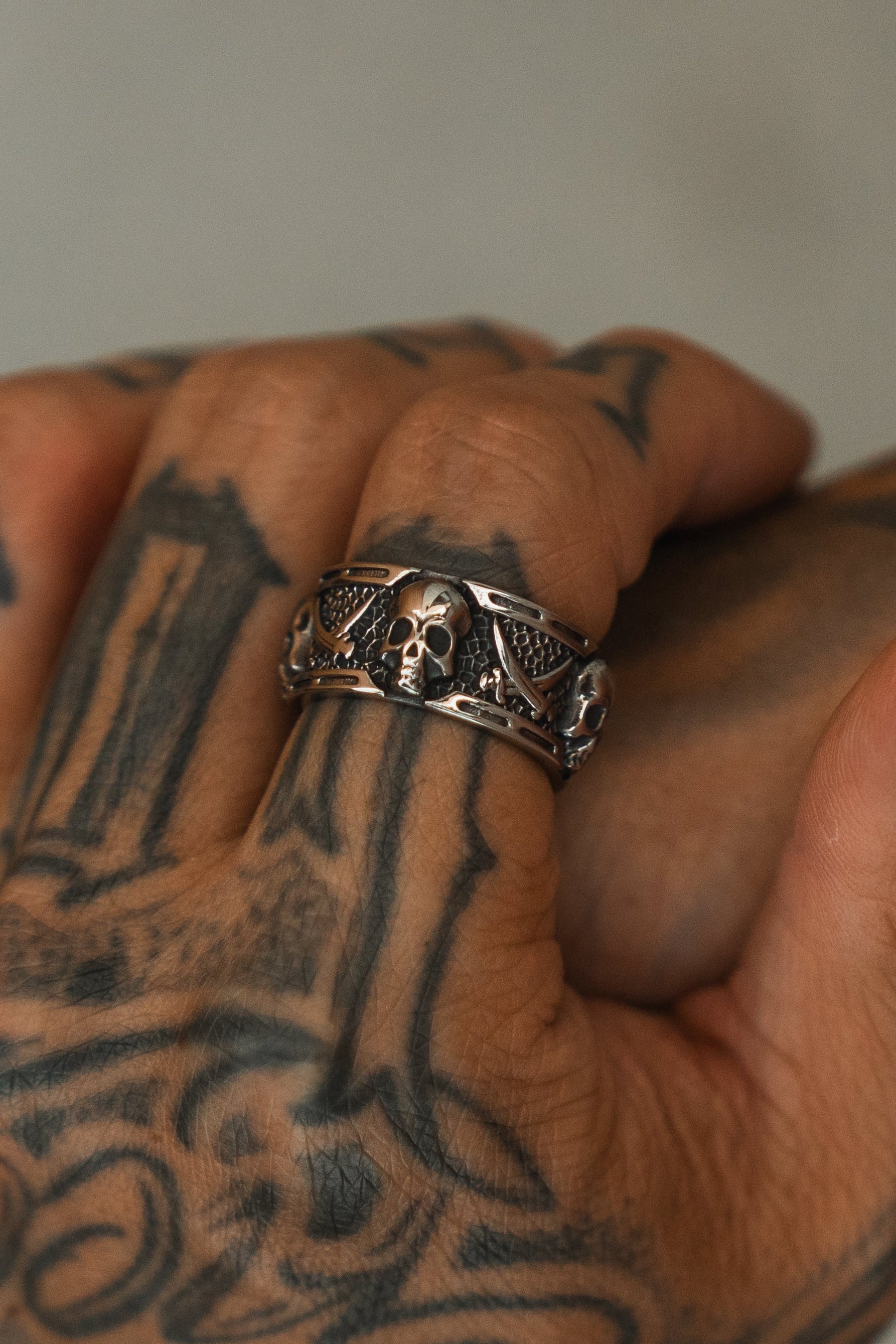 A close-up of a tattooed hand wearing the Relentless Betrayal Piracy Ring, featuring bold skull and wing details. The blurred background highlights the intricate tattoos and the standout ring design.