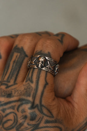 A close-up of a tattooed hand wearing the Relentless Betrayal Piracy Ring, featuring bold skull and wing details. The blurred background highlights the intricate tattoos and the standout ring design.