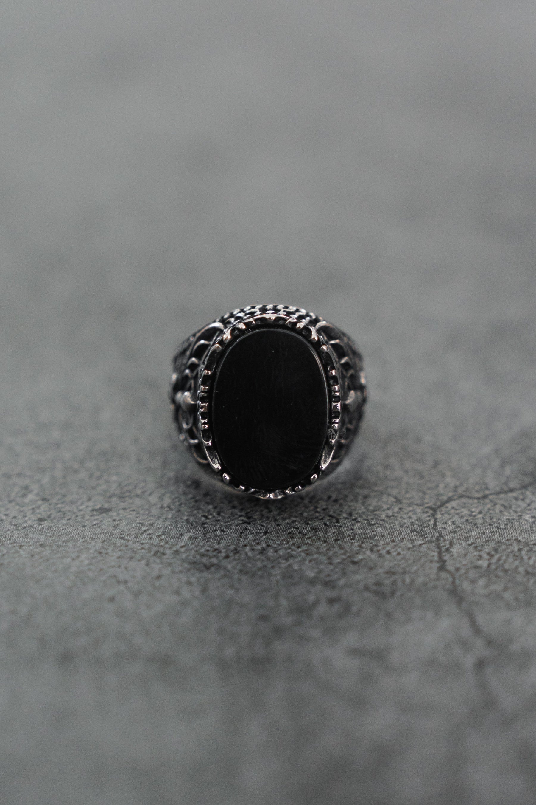 A close-up of the Royal Darkness Ring by Relentless Betrayal, a stainless steel ring with an oval black stone, intricate detailing, and a striking black finish on the band, displayed on a textured gray surface.