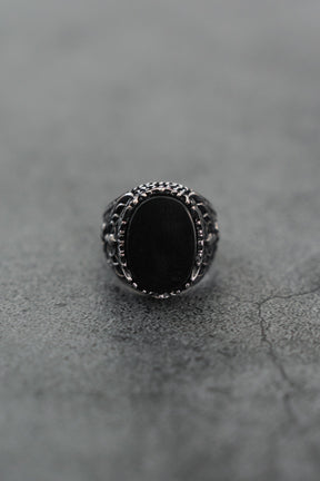 A close-up of the Royal Darkness Ring by Relentless Betrayal, a stainless steel ring with an oval black stone, intricate detailing, and a striking black finish on the band, displayed on a textured gray surface.