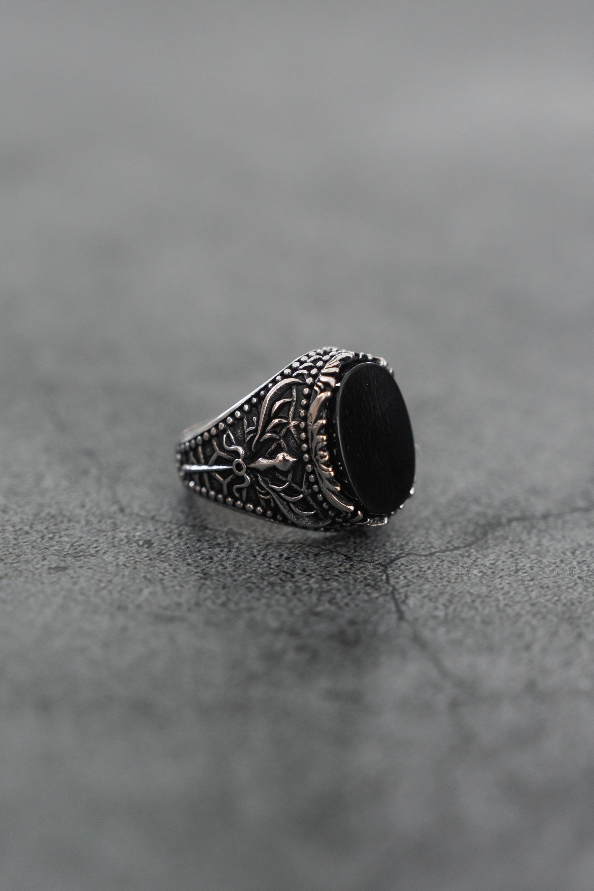 The Dark Current Ring by Relentless Betrayal, a handcrafted men's ring featuring a large black oval stone and intricate detailing, is displayed on a textured gray surface.