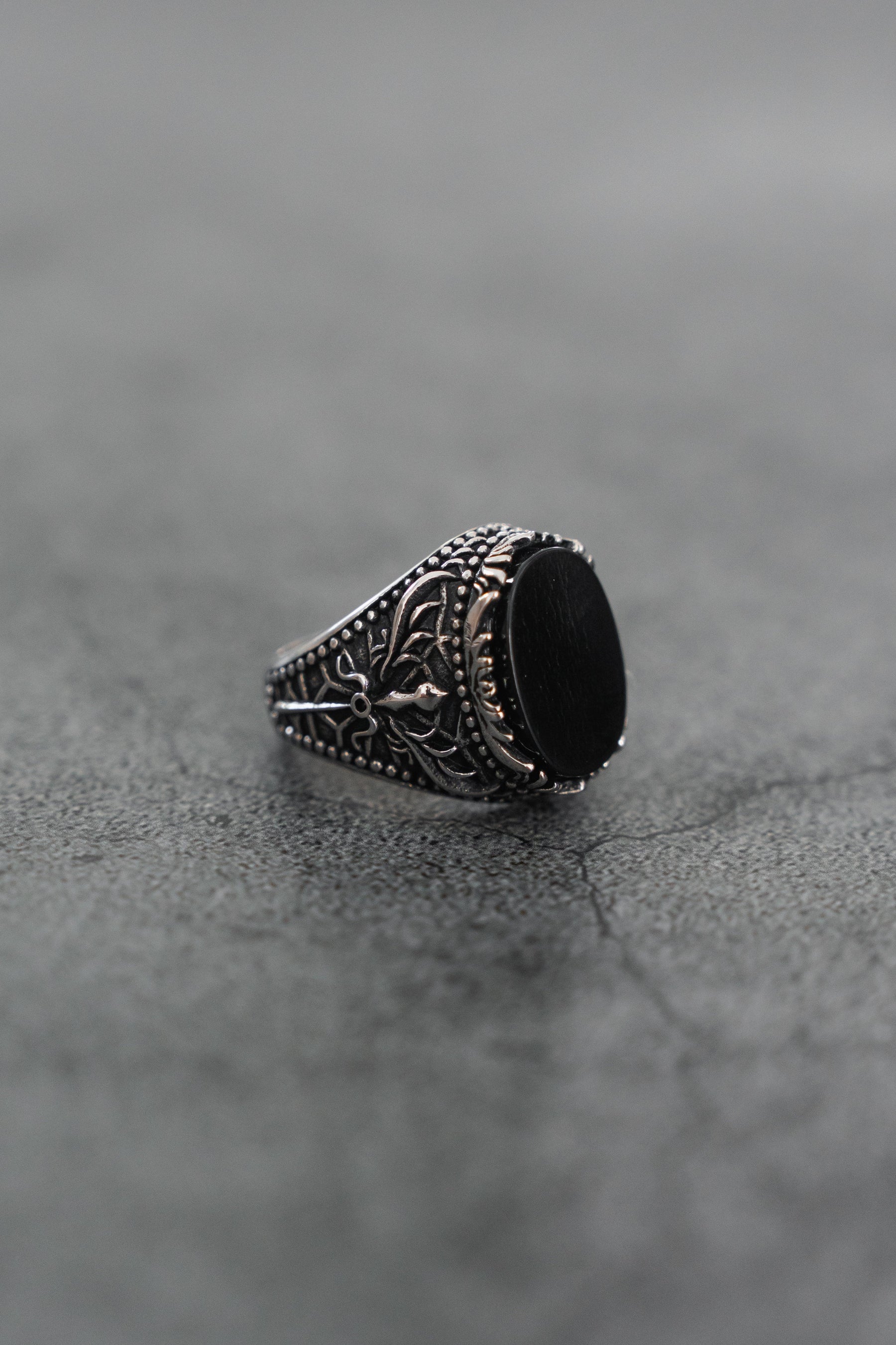 The Dark Current Ring by Relentless Betrayal, a handcrafted men's ring featuring a large black oval stone and intricate detailing, is displayed on a textured gray surface.