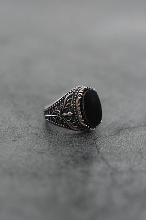 The Dark Current Ring by Relentless Betrayal, a handcrafted men's ring featuring a large black oval stone and intricate detailing, is displayed on a textured gray surface.