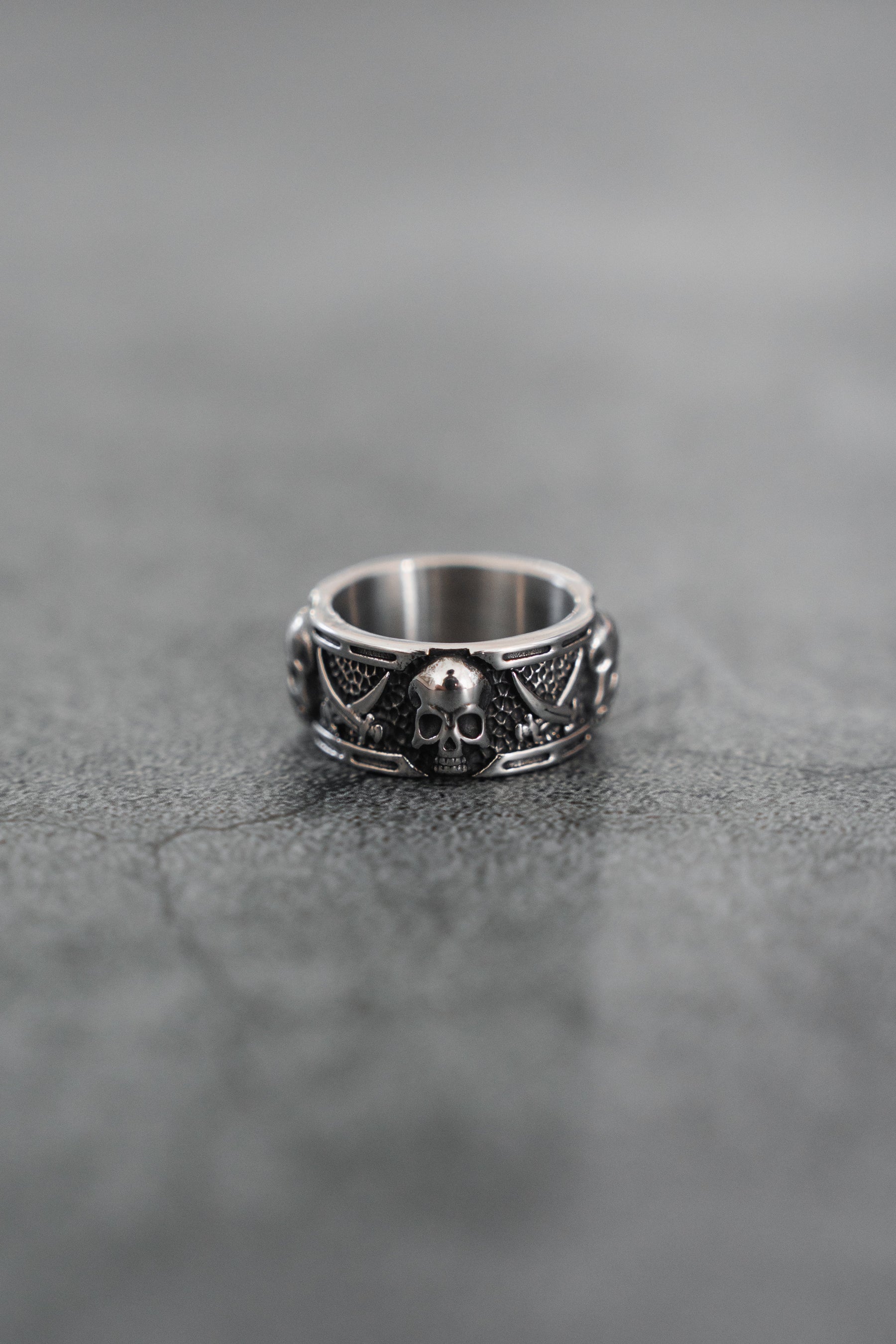 The Piracy Ring by Relentless Betrayal features a bold stainless steel skull on the front, accented by intricate patterns and smaller skulls along the band, all set against a textured gray surface for a striking look.