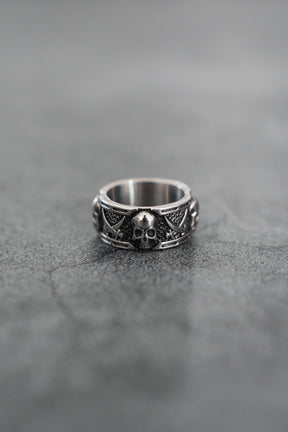 The Piracy Ring by Relentless Betrayal features a bold stainless steel skull on the front, accented by intricate patterns and smaller skulls along the band, all set against a textured gray surface for a striking look.