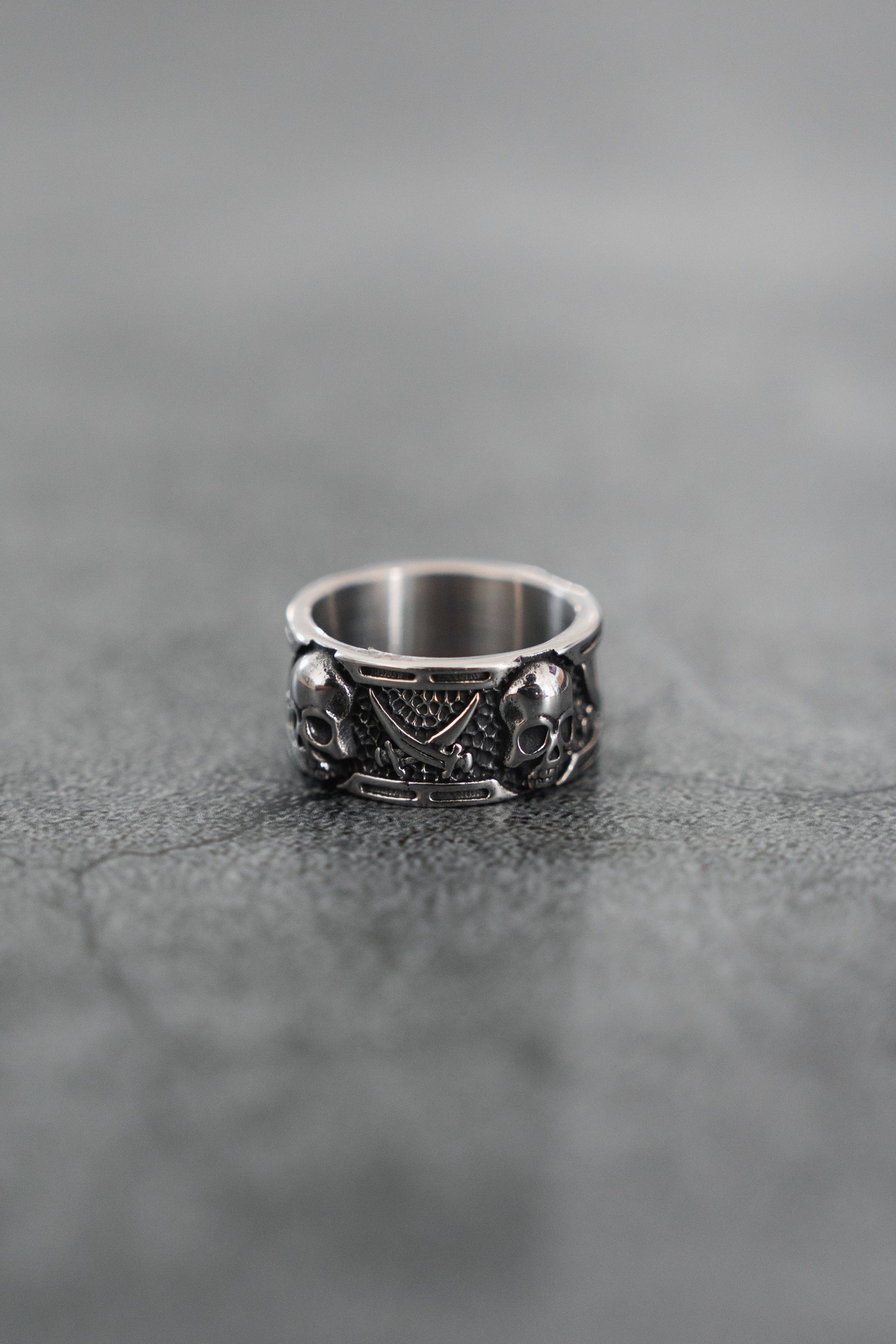 The Piracy Ring by Relentless Betrayal is a stainless steel piece with intricate details, showcasing two skulls and a triangular pattern, displayed on a textured gray surface.