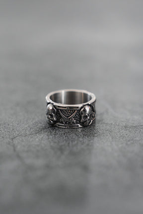 The Piracy Ring by Relentless Betrayal is a stainless steel piece with intricate details, showcasing two skulls and a triangular pattern, displayed on a textured gray surface.