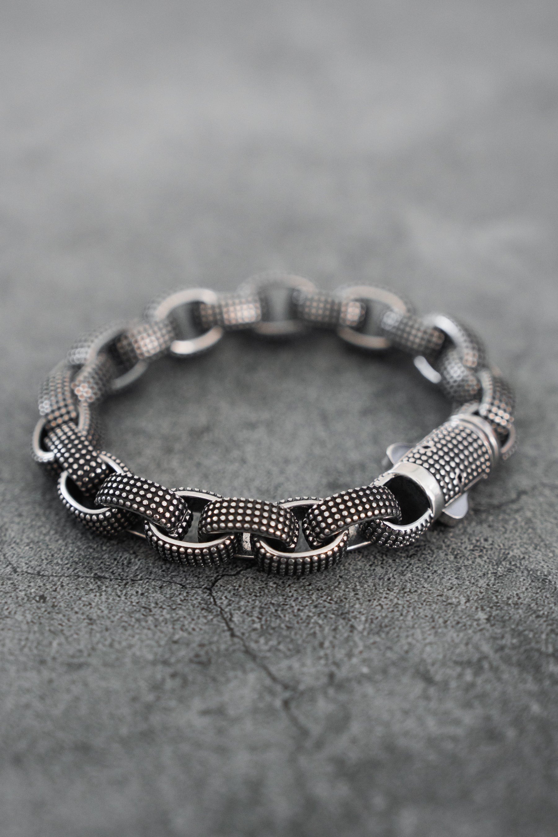 The Relentless Betrayal Seeker Bracelet, hand-forged from stainless steel with large interlocking links and a textured silver-tone finish, is shown on a gray concrete surface.
