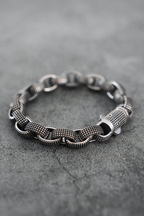 The Relentless Betrayal Seeker Bracelet, hand-forged from stainless steel with large interlocking links and a textured silver-tone finish, is shown on a gray concrete surface.