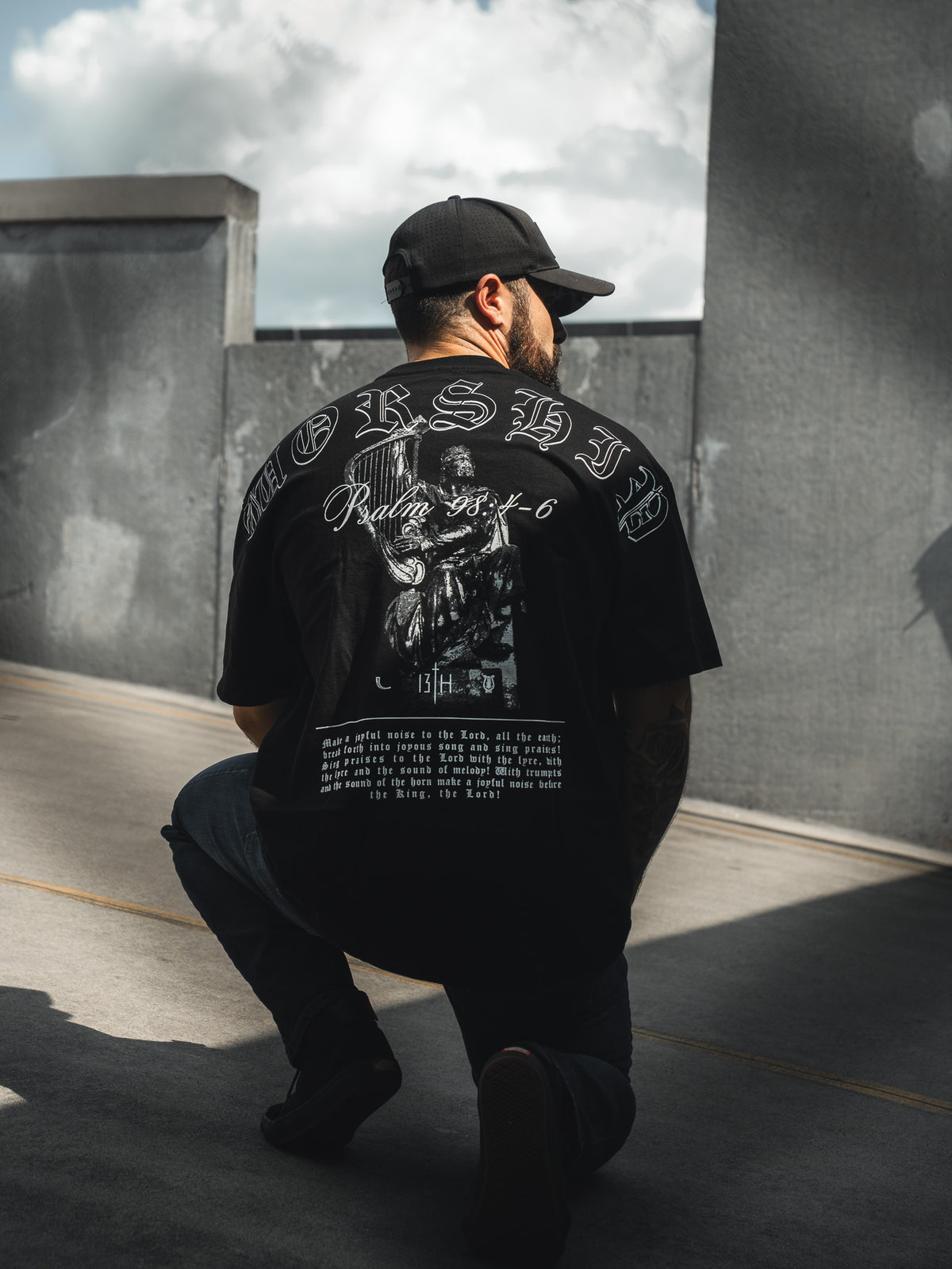 A man wearing the 13th Unwavering Worship Premium Oversized Tee and a black cap kneels on a sunlit rooftop, facing away. Framed by concrete walls under partly cloudy skies, the scene reflects the spirit of Psalm 98:4-6.