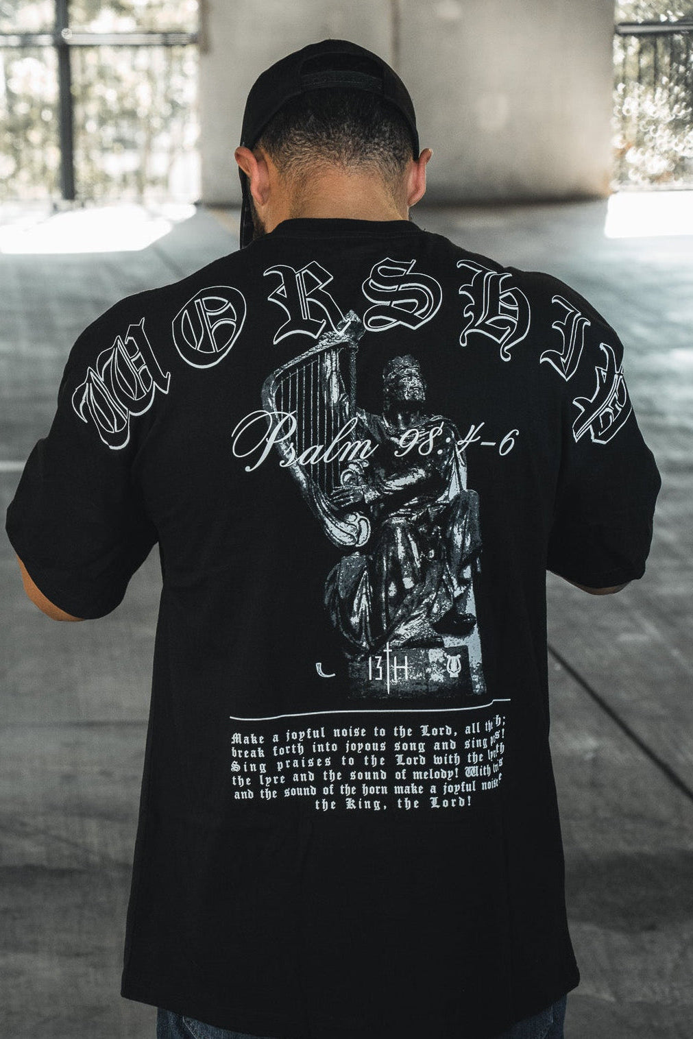 A man faces away in a sunlit concrete space, wearing the 13th Unwavering Worship Premium Oversized Tee featuring “WORSHIP,” “Psalm 138:1-6,” and a praying figure—an expression of devotion inspired by Psalm 98:4-6.
