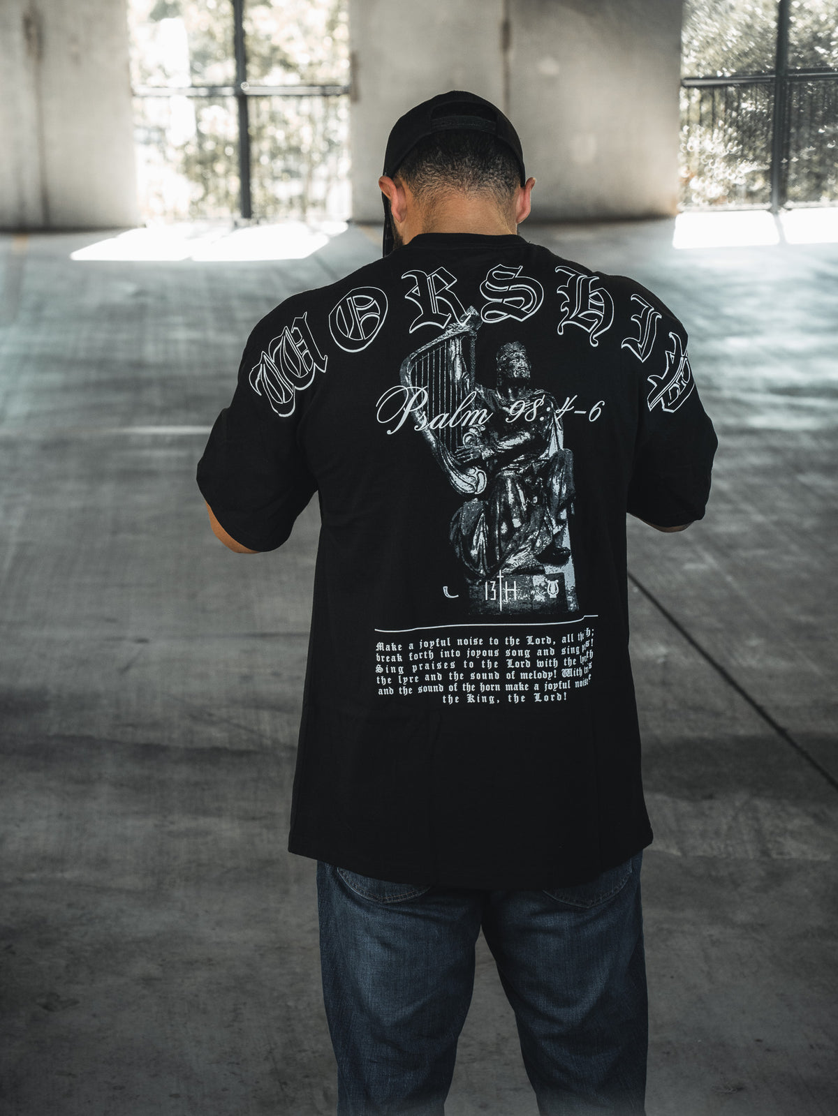 A man faces away in a sunlit concrete space, wearing the 13th Unwavering Worship Premium Oversized Tee featuring “WORSHIP,” “Psalm 138:1-6,” and a praying figure—an expression of devotion inspired by Psalm 98:4-6.