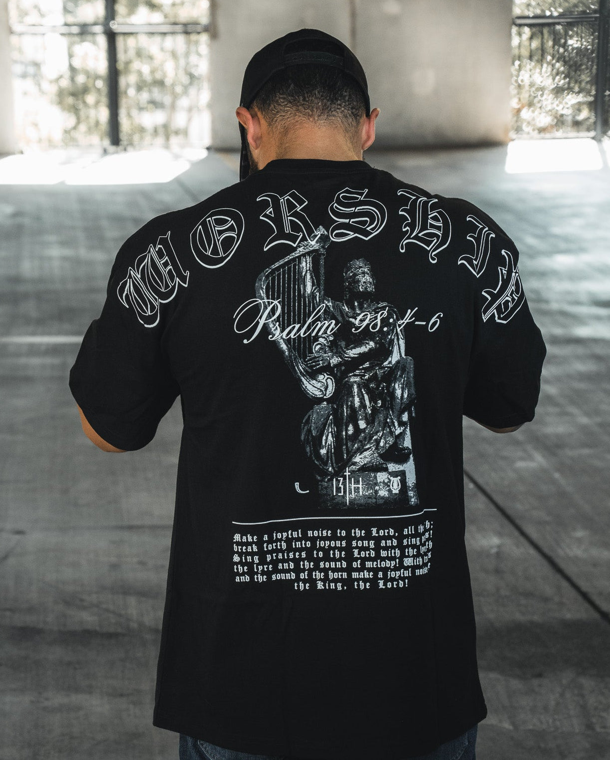 A man faces away in a sunlit concrete space, wearing the 13th Unwavering Worship Premium Oversized Tee featuring “WORSHIP,” “Psalm 138:1-6,” and a praying figure—an expression of devotion inspired by Psalm 98:4-6.