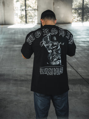 A man faces away in a sunlit concrete space, wearing the 13th Unwavering Worship Premium Oversized Tee featuring “WORSHIP,” “Psalm 138:1-6,” and a praying figure—an expression of devotion inspired by Psalm 98:4-6.