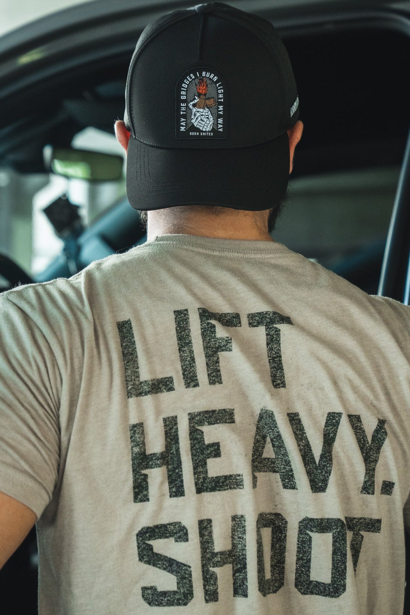 A person in the Born United Premium Snapback - Burning Bridges and a “LIFT HEAVY. SHOOT GUNS.” tee stands by an open car door, seen from behind—ready for anything, embodying the fearless spirit of Born United Private Label.