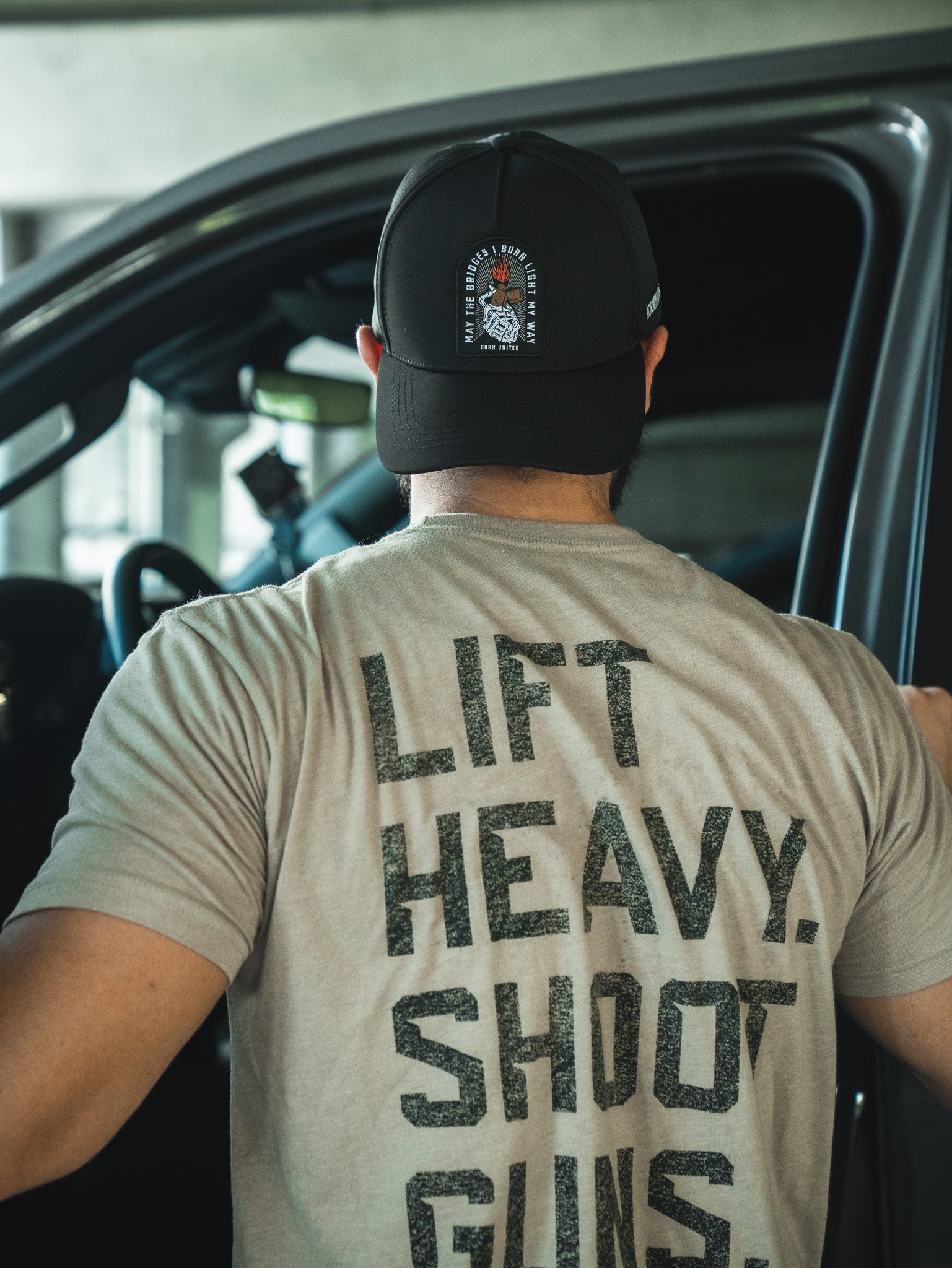 A person in the Born United Premium Snapback - Burning Bridges and a “LIFT HEAVY. SHOOT GUNS.” tee stands by an open car door, seen from behind—ready for anything, embodying the fearless spirit of Born United Private Label.