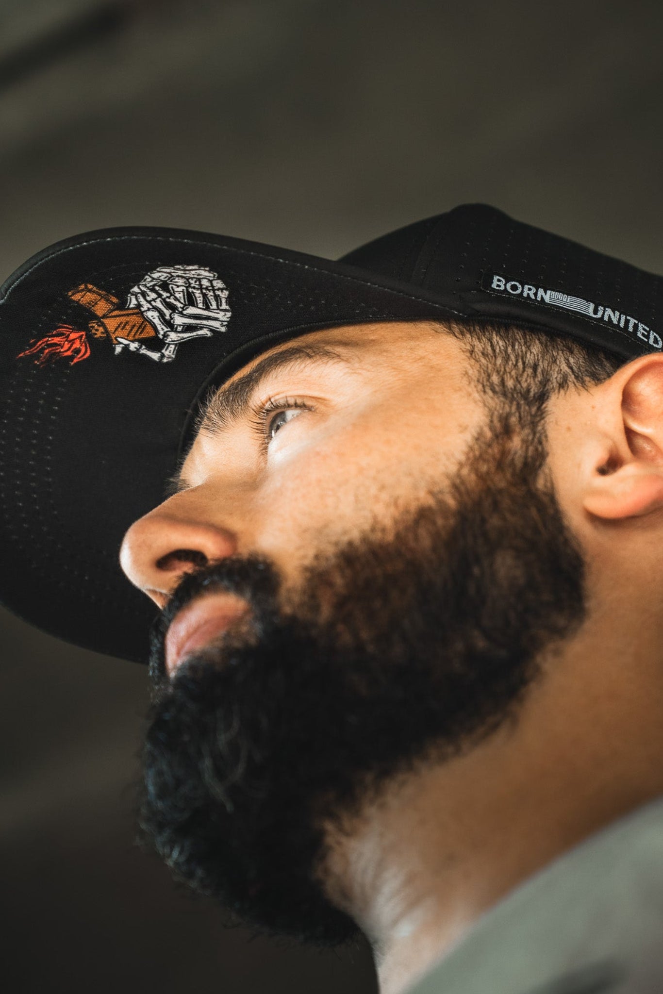 A bearded man wearing the Born United Premium Snapback - Burning Bridges by Born United Private Label, featuring embroidered designs and “BORN UNITED” on the side, looks upward. Subtle "Burning Bridges" details accent the black cap. Shot from a low angle in soft light.