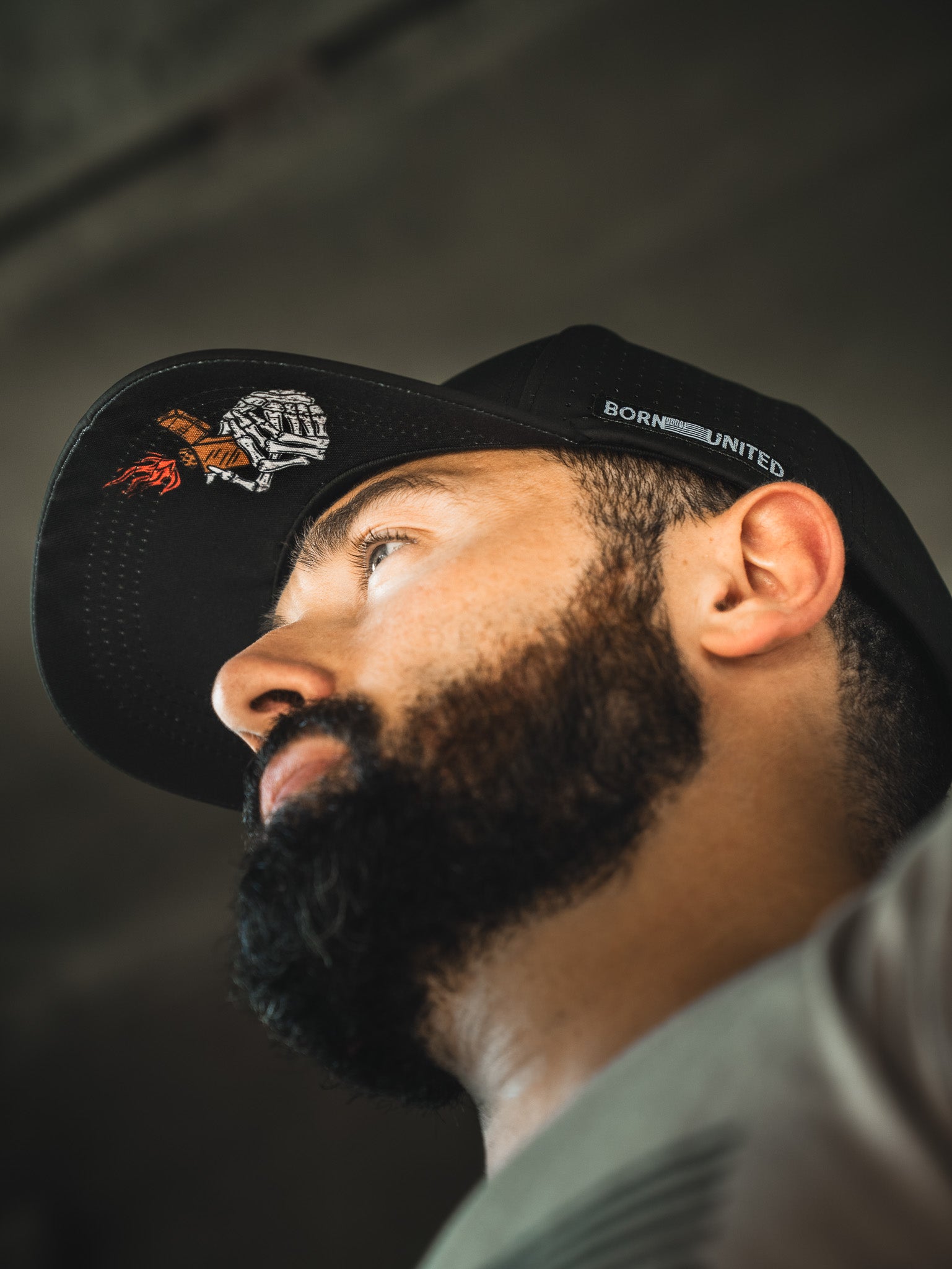 A bearded man wearing the Born United Premium Snapback - Burning Bridges by Born United Private Label, featuring embroidered designs and “BORN UNITED” on the side, looks upward. Subtle "Burning Bridges" details accent the black cap. Shot from a low angle in soft light.