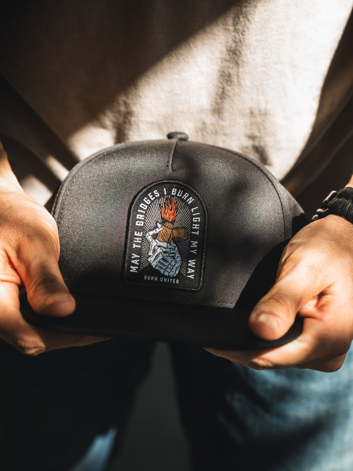 A person holds the Born United Premium Snapback - Burning Bridges by Born United Private Label, featuring a flaming hand patch and the quote "May the bridges I burn light my way," highlighted in sunlight to symbolize bold new paths.