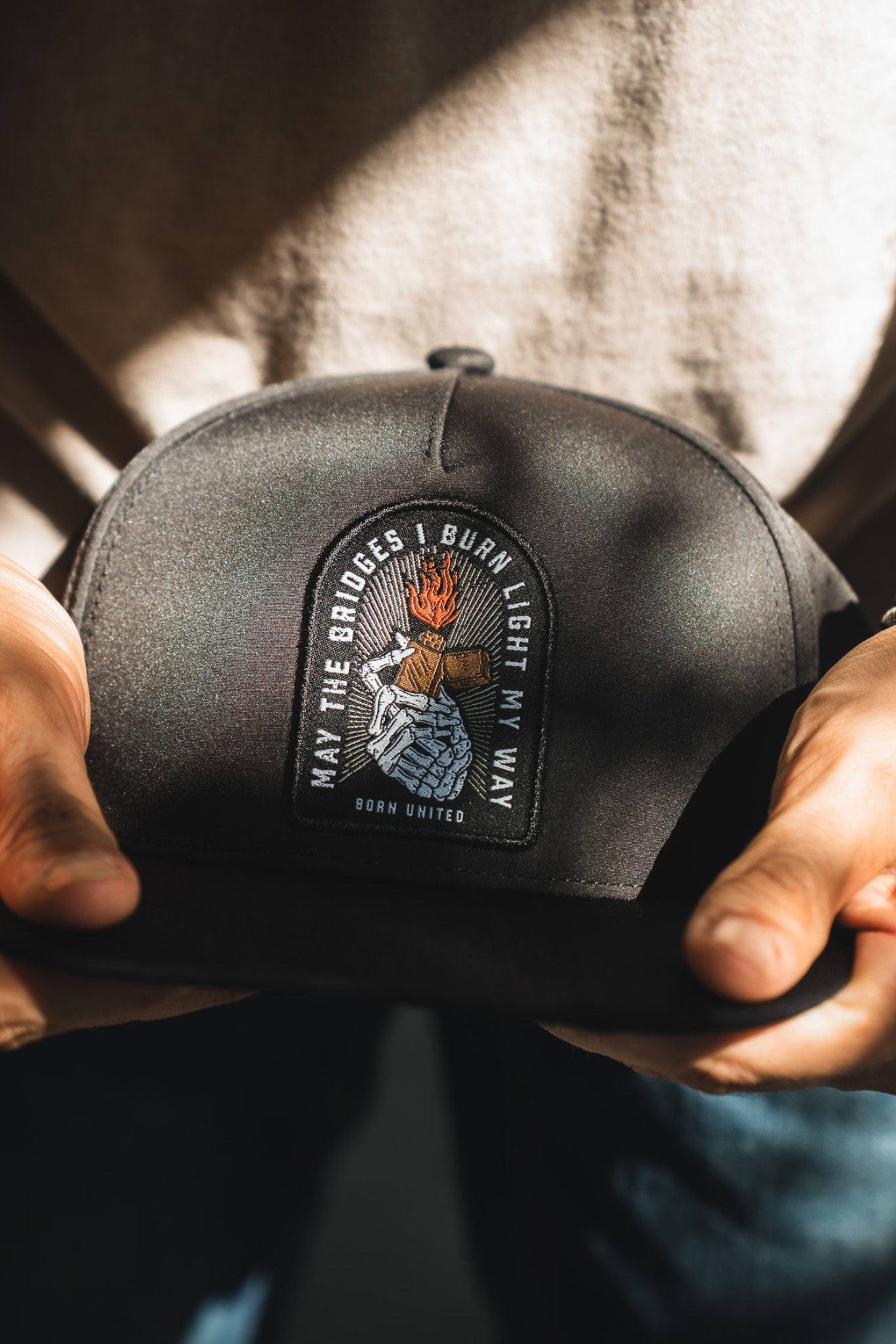 A person holds the Born United Premium Snapback - Burning Bridges by Born United Private Label, featuring a flaming hand patch and the quote "May the bridges I burn light my way," highlighted in sunlight to symbolize bold new paths.