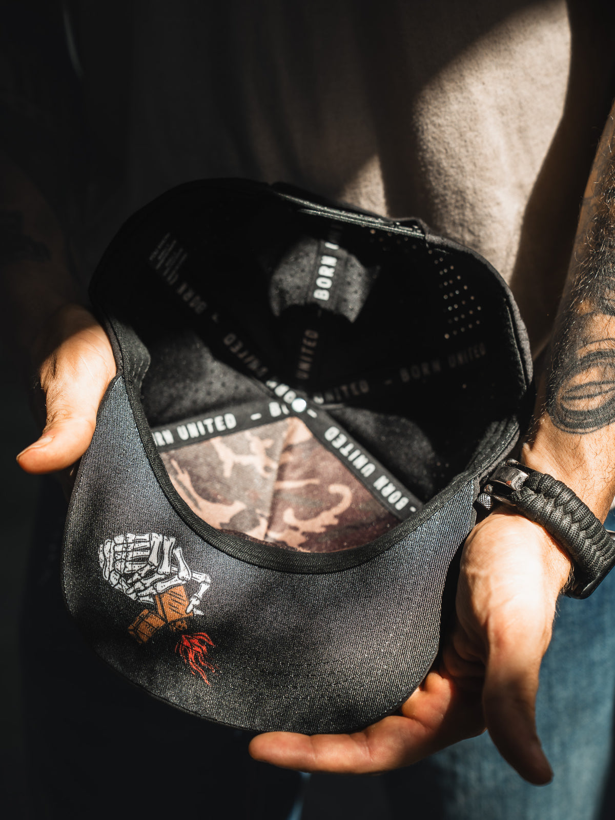A person holds open the Born United Premium Snapback - Burning Bridges by Born United Private Label, revealing a skeleton hand with money and a rose on the brim, plus "Burning Bridges" and "BORN UNITED" text on the inner band.