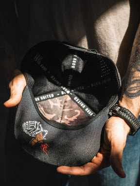 A person holds open the Born United Premium Snapback - Burning Bridges by Born United Private Label, revealing a skeleton hand with money and a rose on the brim, plus "Burning Bridges" and "BORN UNITED" text on the inner band.