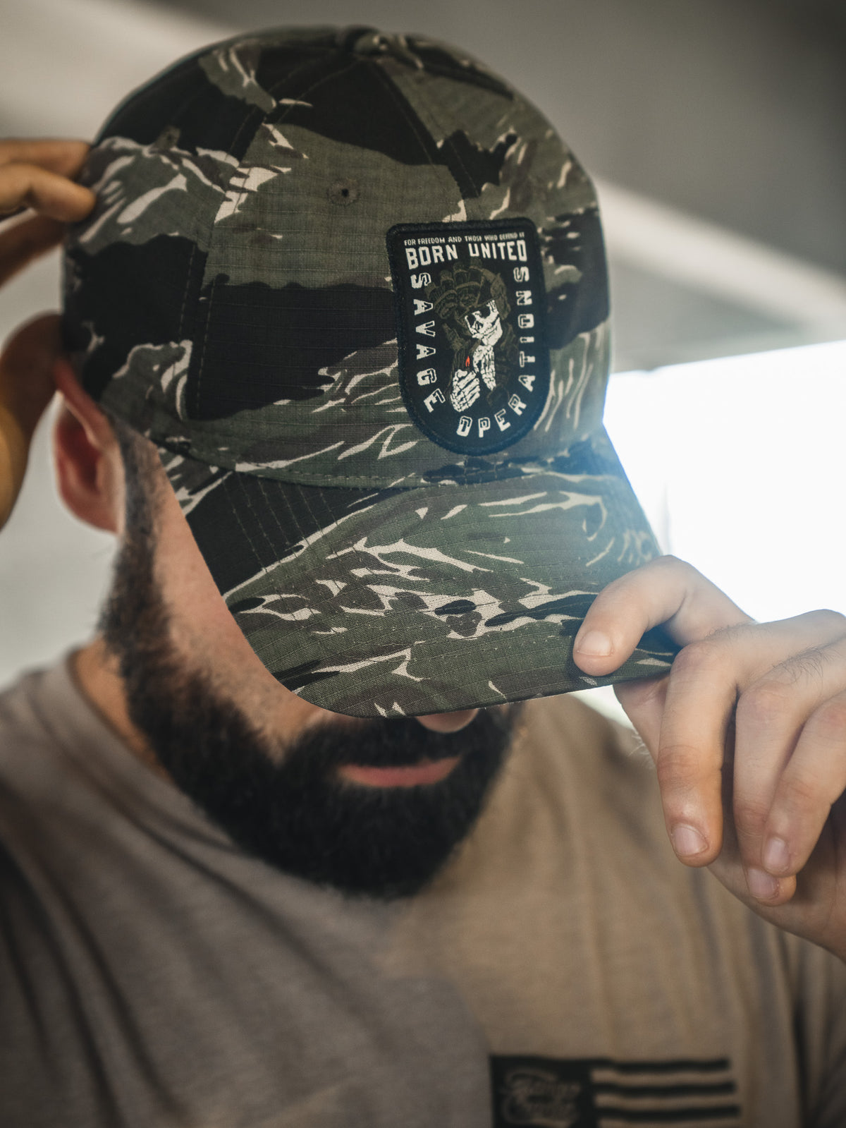 A bearded man in a camouflage Born United Private Label Buckle Hat – Savage Operations, holds the brim and partly obscures his face.
