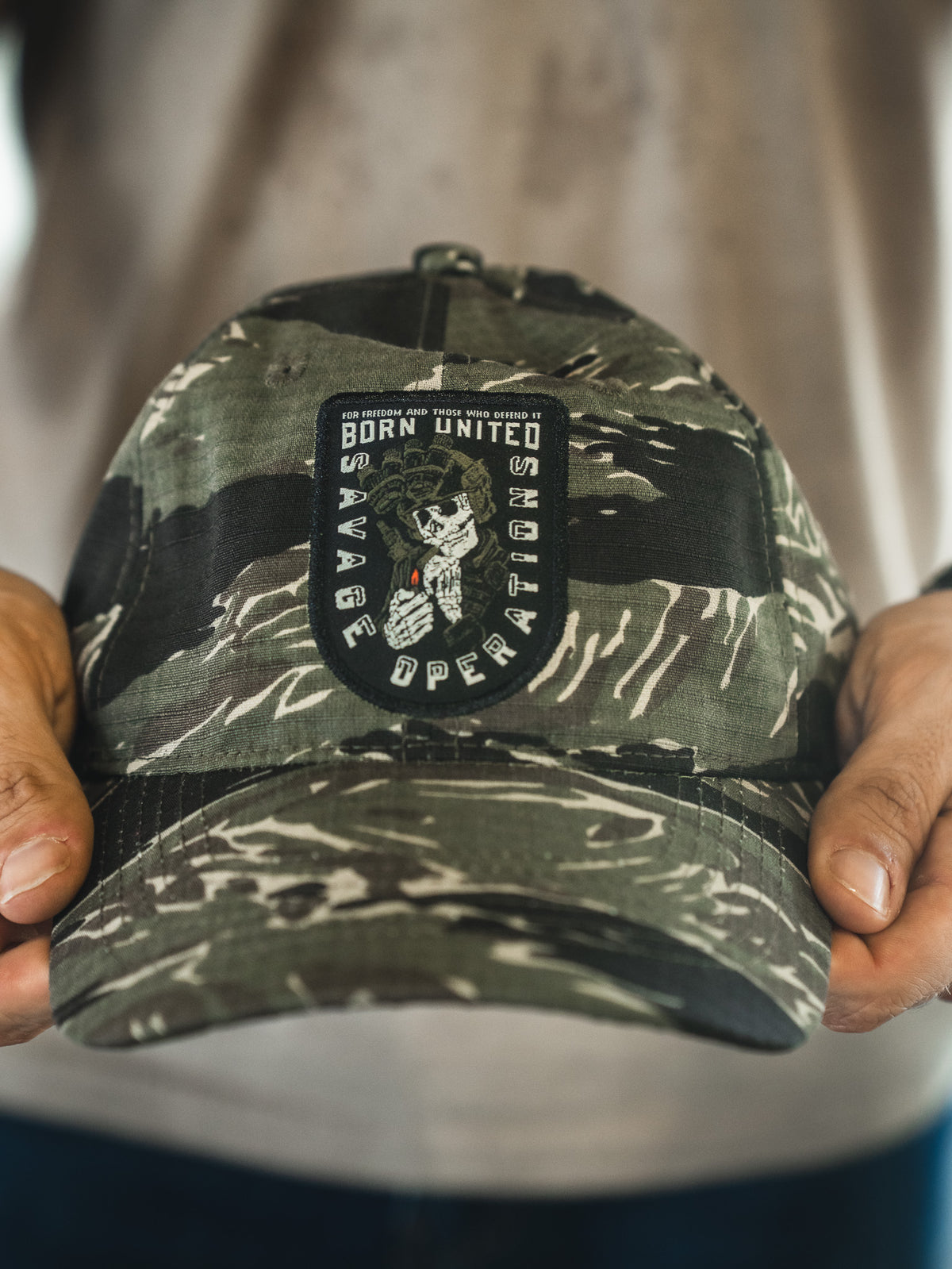A person holds the Born United Buckle Hat - Savage Operations by Born United Private Label, a camouflage cap featuring a patch with "Born United Savage Operations" and an image of a skeleton holding a rifle.