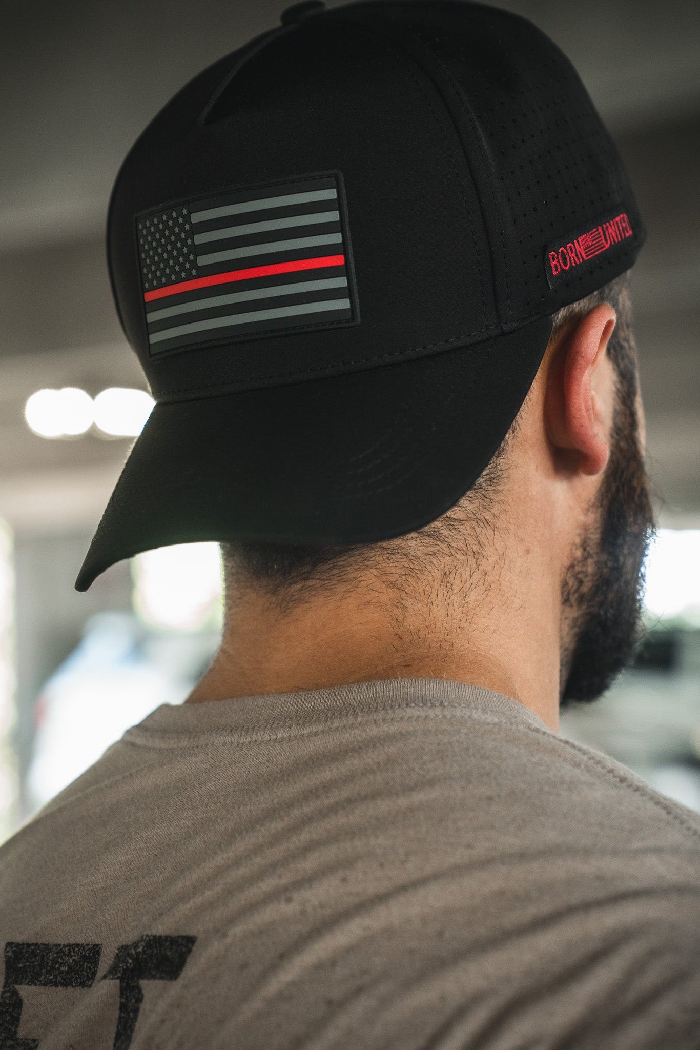 A man in a light gray shirt is seen from behind wearing the Born United Premium Flag Snapback - TRL by Born United Private Label, featuring a black cap with a subdued flag patch, red stripe, and "BORN PRIMITIVE" on the side.