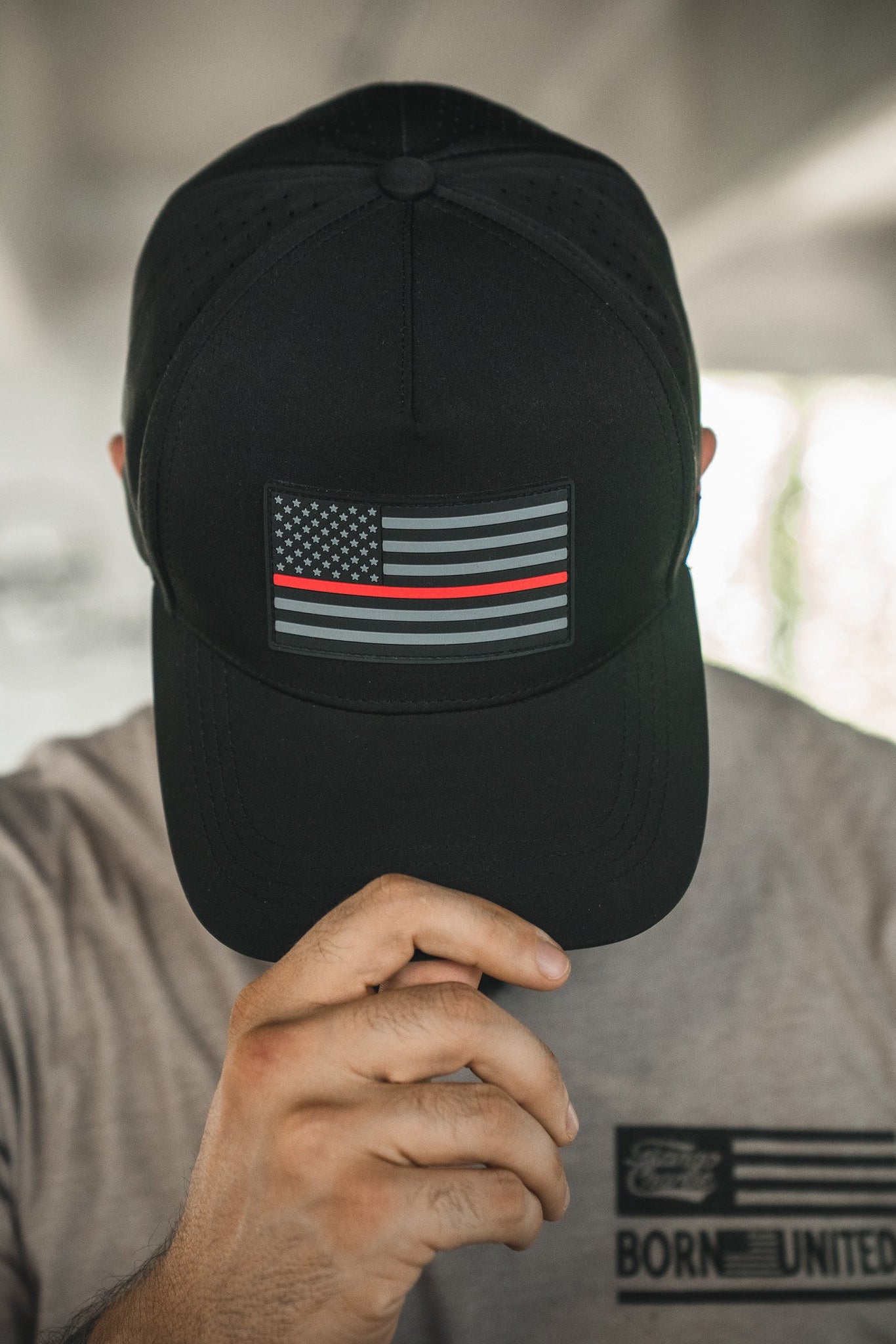 A person wearing the Born United Premium Flag Snapback - TRL by Born United Private Label, featuring a subdued American flag with a thin red line, partially covering their face, and a gray shirt with "Born United" printed on it.