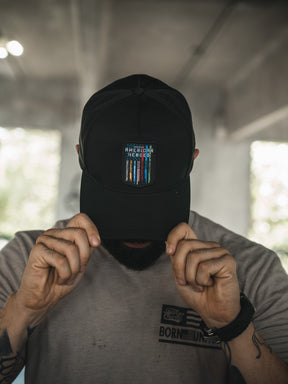 A person in a light gray t-shirt and black cap holds the brim, facing down. The Born United Private Label cap features a small patch with multicolored lines and text reading "American Heroes" from the Born United Premium Snapback line.