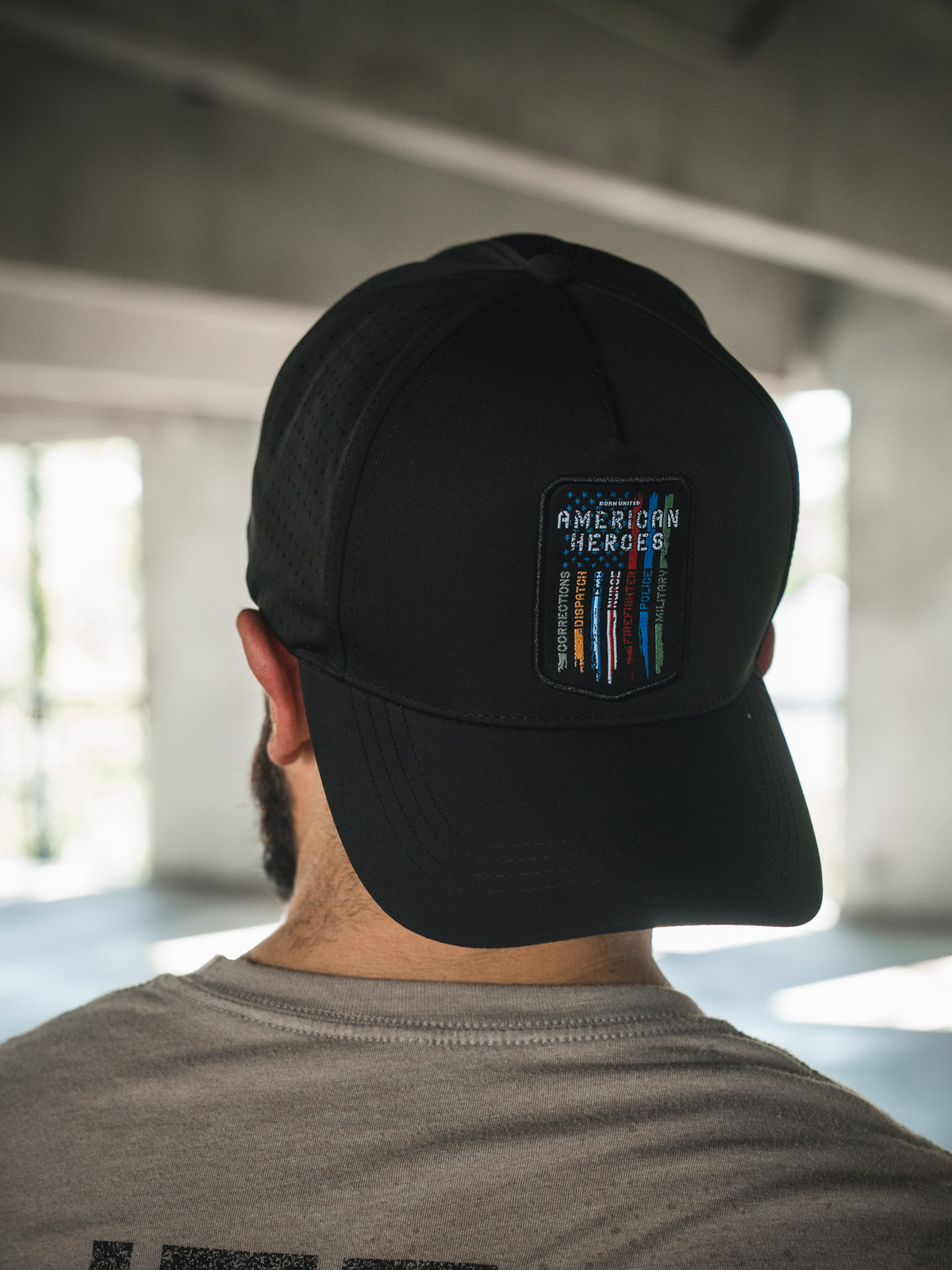 A person seen from behind wearing the Born United Private Label’s Born United Premium Snapback – American Heroes, a black cap with a patch that reads "American Heroes" and colorful vertical lines, indoors with natural light coming through windows.