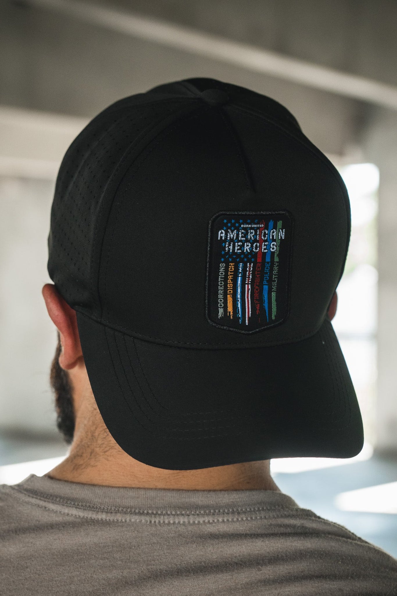 A person seen from behind wearing the Born United Private Label’s Born United Premium Snapback – American Heroes, a black cap with a patch that reads "American Heroes" and colorful vertical lines, indoors with natural light coming through windows.
