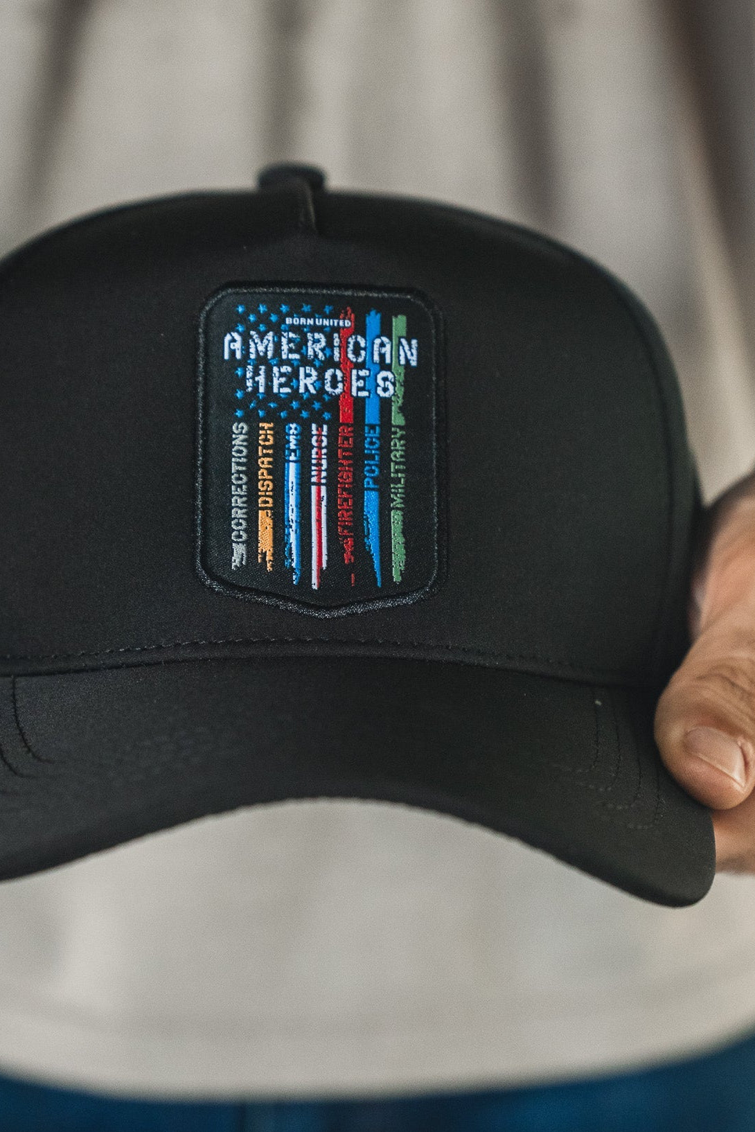 A person holds the Born United Premium Snapback - American Heroes by Born United Private Label, featuring a black cap with a colorful "AMERICAN HEROES" patch and vertical red, white, blue, and green stripes. The wearer has a light shirt and blue jeans.