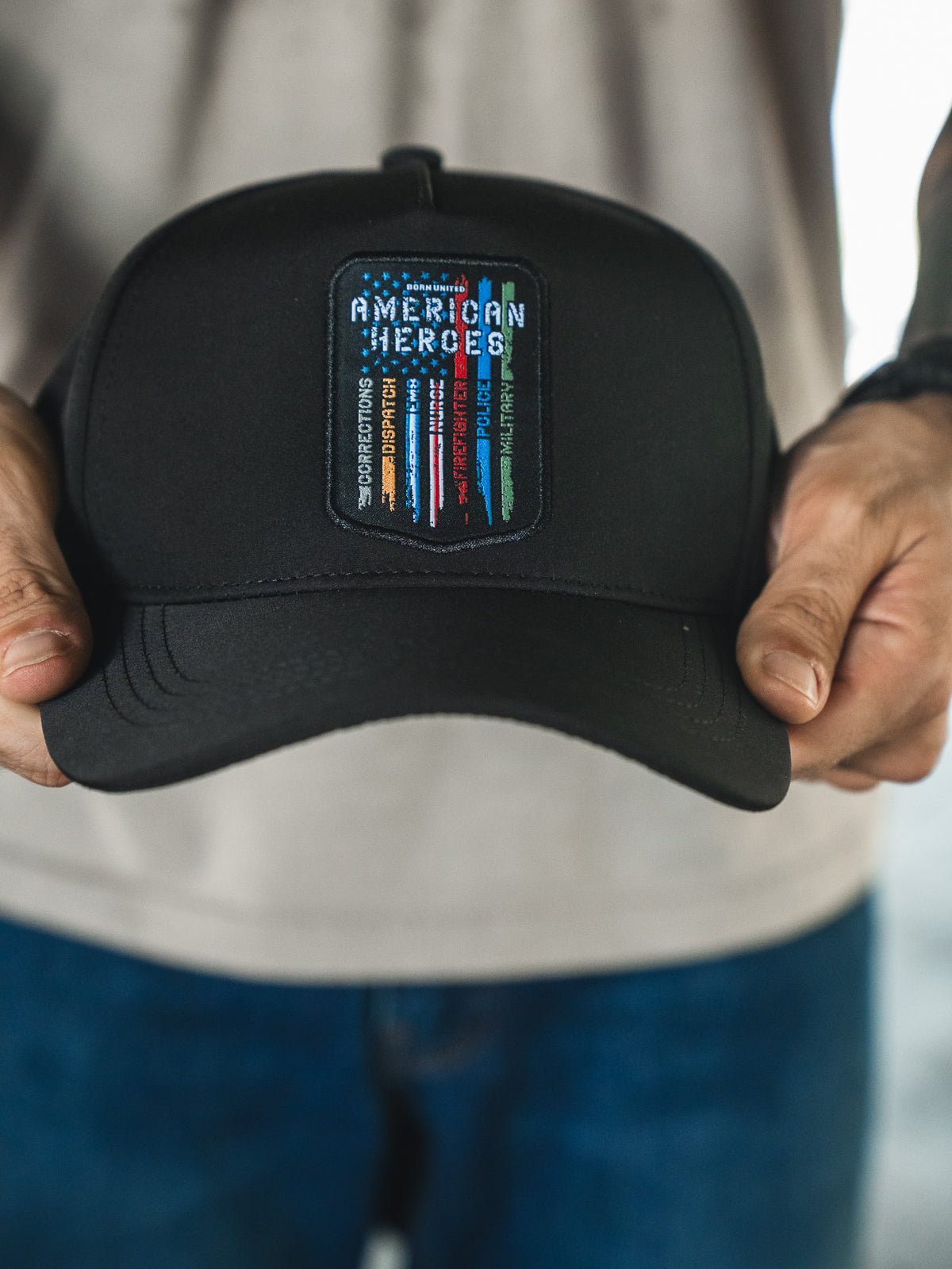 A person holds the Born United Premium Snapback - American Heroes by Born United Private Label, featuring a black cap with a colorful "AMERICAN HEROES" patch and vertical red, white, blue, and green stripes. The wearer has a light shirt and blue jeans.